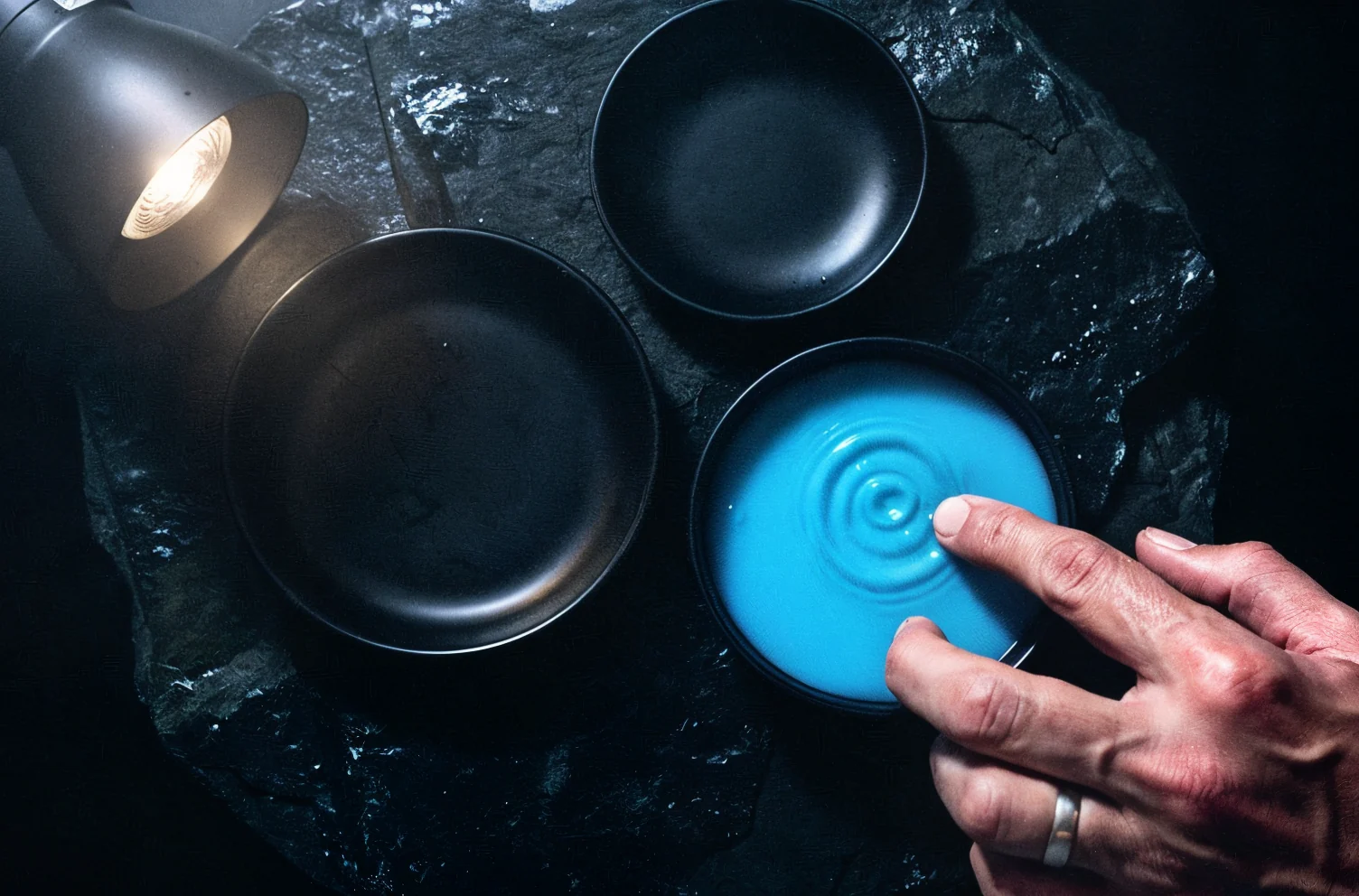 Overhead view of four black dishes set into wet stone, one filled with glowing blue liquid and a hand touching its surface.
