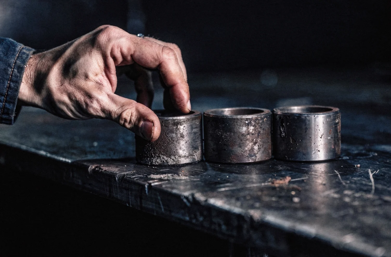 A hand setting a worn steel component at the end of a short row of four similar components laid out with even spacing on a dark steel bench