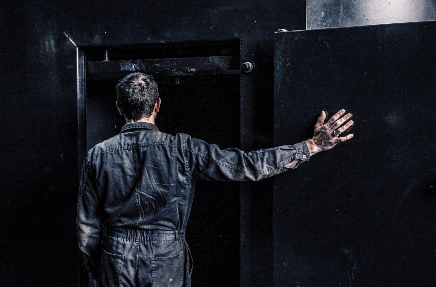 A person in creased dark work clothes seen from behind, one hand flat on the open steel door of a large dark plant room