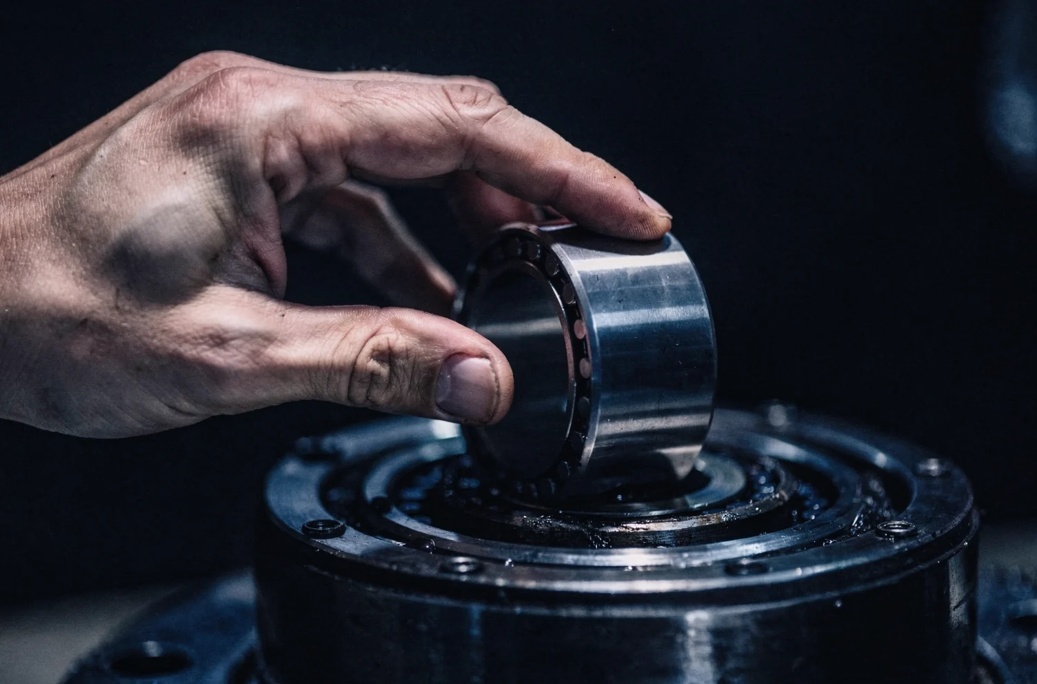One hand lifting a stainless steel roller bearing out of a greased circular seat in a dark machined steel housing.