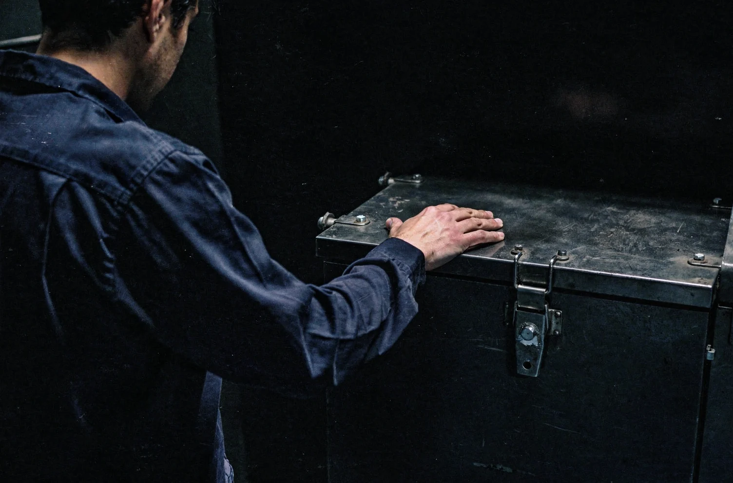 A person in a creased navy work shirt seen from behind, one hand resting flat on the closed lid of a heavy steel case.