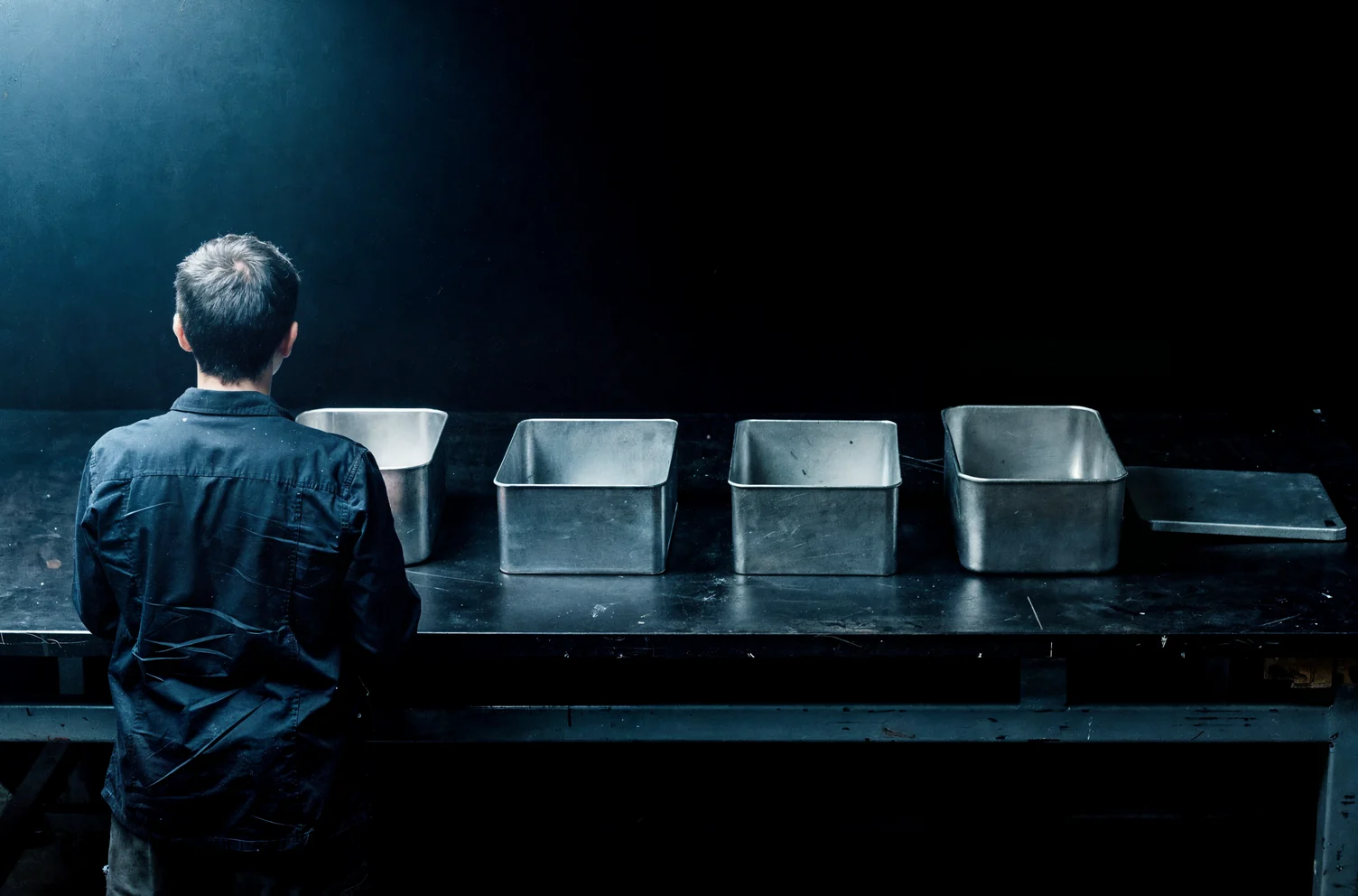 One person seen from behind standing at a dark steel bench in front of four plain steel vessels in a row, all open and empty